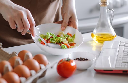 Woman preparing diet salad using digital cookbook. Fresh vegetables and laptop in home kitchen. Online cooking. Record recipes on camera.